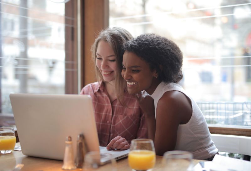 Two friends smiling together at a laptop in a cafe