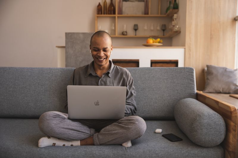 A man laughing as he uses a MacBook on a sofa at home